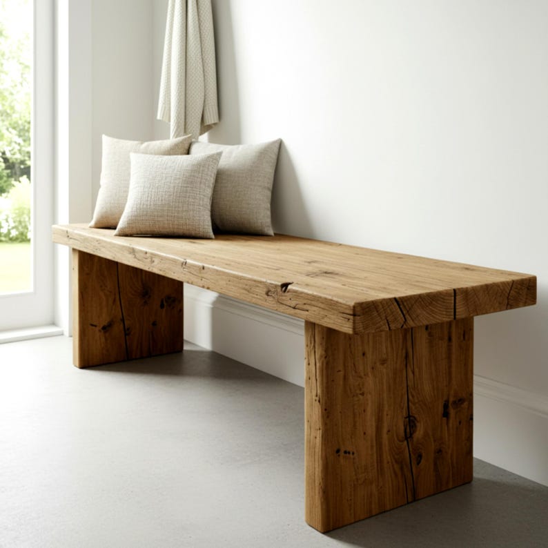 Puede incluir: Banco de madera maciza con acabado natural. El banco tiene un asiento rectangular y patas gruesas en forma de bloque. Tres cojines cuadrados de color neutro descansan sobre el banco. El banco mide aproximadamente 152 cm de largo.