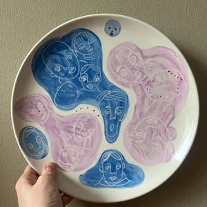 Könnte beinhalten: Ein weißer Keramikteller mit einem handgemalten Design mit abstrakten Gesichtern in Blau- und Lavendeltönen. Die Gesichter sind weiß umrandet, mit ein paar kleinen roten Punkten. Der Teller hat eine runde Form.