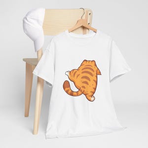 May include: White t-shirt featuring a cartoon cat with orange and brown stripes. The cat is depicted from the back, with its tail curled. The shirt is hanging on a wooden hanger, with a white baseball cap resting on a wooden chair.