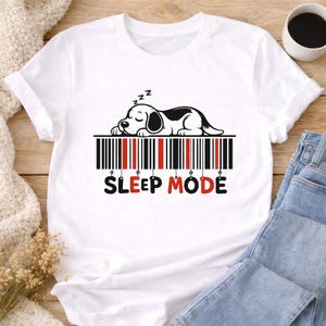 Puede incluir: Camiseta blanca con un diseño de un perro durmiendo sobre un código de barras. El texto "SLEEP MODE" está en negro y rojo. El perro es blanco y negro, con los ojos cerrados y símbolos "z" sobre su cabeza.