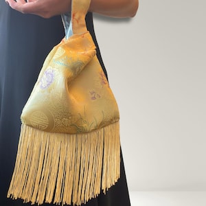 Peut inclure: Un sac poignet jaune en tissu à motifs, orné de fleurs et de symboles géométriques. Ce sac dispose d'une anse unique et se termine par de longues franges fines de couleur jaune assortie, suspendues au bord inférieur de la pochette.