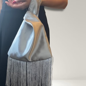 May include: A silver satin wristlet handbag featuring a triangular silhouette with a single integrated handle and a long, flowing fringe trim along the bottom edge. The smooth, lustrous fabric creates soft folds, while the delicate fringe adds texture.
