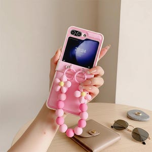 May include: Pink phone case with a flower and bead strap. The case is holding a phone with a screen displaying the time 21:24. The strap features pink flowers and pink beads. The phone case is on a wooden table.