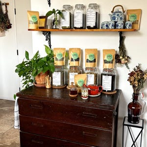 May include: A display of tea and tea accessories. A dark wooden dresser holds jars of tea, tea bags, a small red teapot, and a small glass jar. Shelves above hold more tea, teacups, and a red teapot.