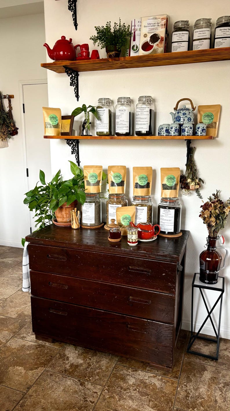 May include: A display of tea and tea accessories. A dark wooden dresser holds jars of tea, tea bags, a small red teapot, and a small glass jar. Shelves above hold more tea, teacups, and a red teapot.