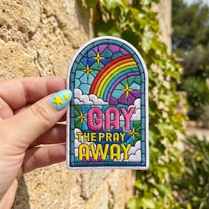 May include: Embroidered patch with a stained-glass window design. The patch features a rainbow, stars, and the words "GAY THE PRAY AWAY" in turquoise and yellow. The patch has a blue border and is held by a hand with a blue nail.