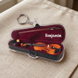 May include: A clear acrylic keychain featuring a miniature violin in its case. The violin is a warm brown color, and the case is black with a burgundy interior. The name "Benjamin" is printed on the case.