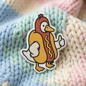 May include: An embroidered patch featuring a white duck wearing a hot dog costume. The duck has a yellow beak, orange feet, and is giving a thumbs-up gesture. The hot dog costume is brown with a yellow mustard squiggle down the center.