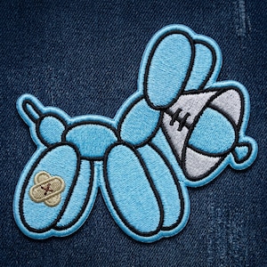 May include: Embroidered patch featuring a light blue balloon dog design with black outlines. The dog has a bandage on one leg and a gray and white football-shaped object. The patch is on a dark blue denim background.