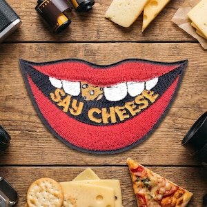 May include: Embroidered patch in the shape of a smiling mouth with red lips, white teeth, and the words "SAY CHEESE" in yellow. The patch has a black border and is surrounded by cheese, crackers, and camera equipment on a wooden surface.