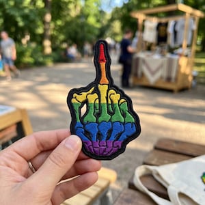 May include: Embroidered patch in the shape of a hand giving the middle finger, with a rainbow gradient design. The patch has a black border and is held up in front of a blurred outdoor background.