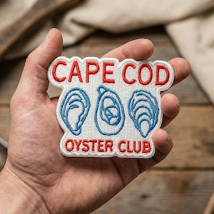 May include: A white embroidered patch with the words "CAPE COD OYSTER CLUB" in red and blue. The patch features three blue oyster illustrations. The patch is held in a hand.