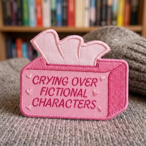 May include: A pink embroidered patch shaped like a tissue box with the text "CRYING OVER FICTIONAL CHARACTERS." The patch has a light pink tissue design coming out of the top and small heart details.