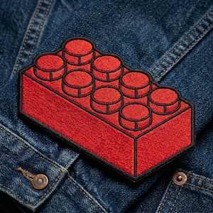 May include: A red embroidered patch in the shape of a classic building block, with black outlining. The patch is sewn onto a blue denim jacket. The patch features seven raised circular studs.
