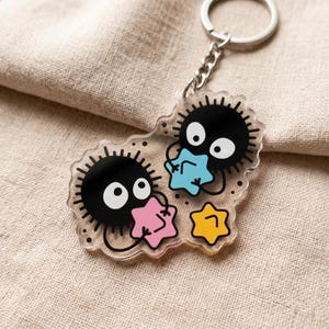 Puede incluir: Llavero de acr&iacute;lico transparente con dos duendes de holl&iacute;n negros con ojos blancos y contornos puntiagudos. Cada duende sostiene una estrella de colores: rosa, azul y amarillo. El llavero tiene una cadena y un anillo plateados.