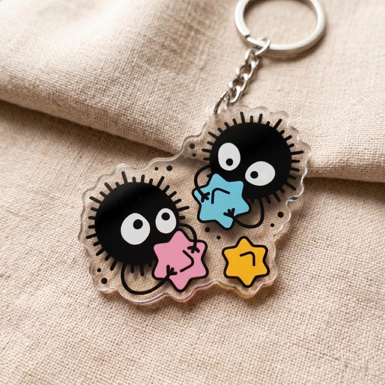 Puede incluir: Llavero de acr&iacute;lico transparente con dos duendes de holl&iacute;n negros con ojos blancos y contornos puntiagudos. Cada duende sostiene una estrella de colores: rosa, azul y amarillo. El llavero tiene una cadena y un anillo plateados.