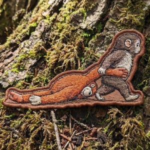 May include: Embroidered patch featuring two monkeys. One monkey is lying down, while the other is walking and holding the other monkey's arm. The patch has a brown border and is set against a background of moss and tree bark.