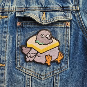 May include: A denim jacket with a patch of a cartoon pigeon wearing a sandwich. The patch features a gray pigeon with orange feet and a yellow and white sandwich around its neck. The jacket is a medium wash blue.