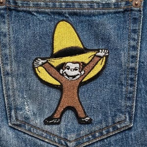 May include: Embroidered patch featuring a cartoon monkey wearing a yellow sombrero. The monkey is brown with black accents, and the patch is set against a blue denim background, likely a pocket. The patch is a fun, playful design.