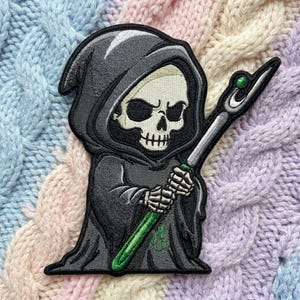 May include: An embroidered patch featuring a grim reaper character wearing a dark gray hooded robe. The figure has a cream-colored skull face and holds a staff with a green gem at the top. The design includes black outlines and green accents.