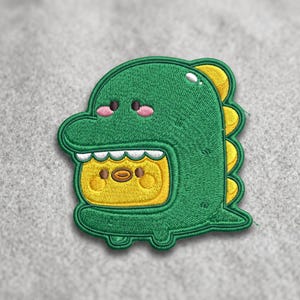 May include: Embroidered patch featuring a green dinosaur costume with yellow accents. The dinosaur has pink cheeks and a yellow creature inside its mouth. The patch is on a gray background.
