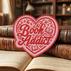 May include: A heart-shaped patch with the words "Book Addict" in red script. The patch is pink with red embroidered details, including stars and small open book illustrations. The patch is placed on top of an open book and other antique books.