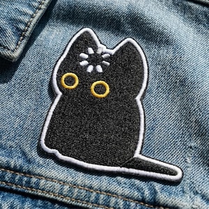 May include: Black cat patch with yellow eyes and a white flower design on its head. The cat patch is outlined in white and is sewn onto a blue denim jacket.