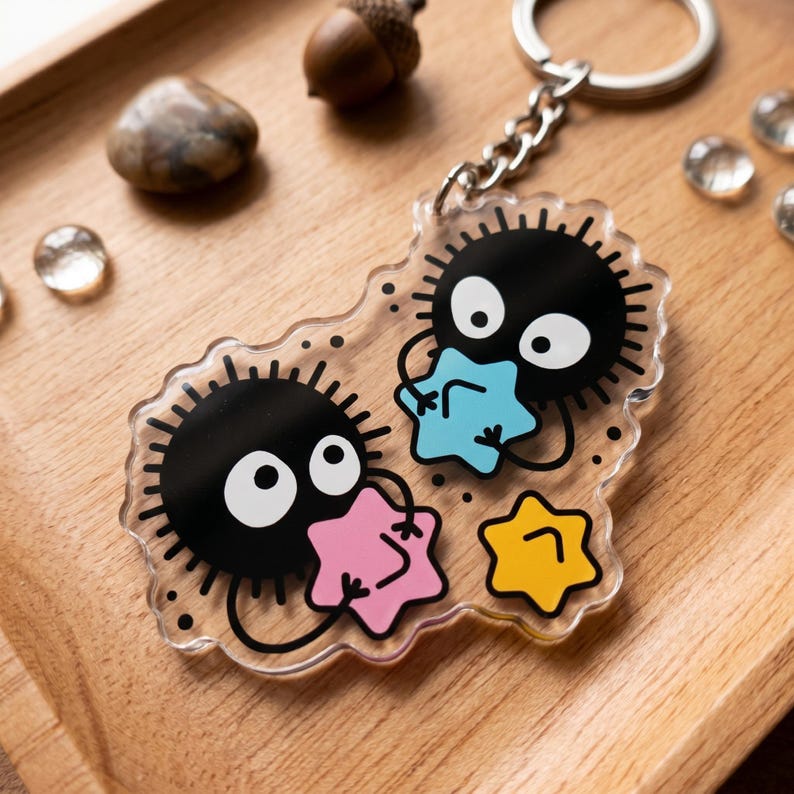 Puede incluir: Un llavero de acr&iacute;lico transparente con dos personajes negros y redondos con ojos blancos que sostienen estrellas de colores. El llavero tiene una cadena y un anillo plateados, sobre un fondo de madera.