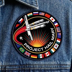 May include: A circular patch with a black background featuring a white spaceship design with red and yellow accents. The patch is surrounded by flags of various countries and the text "PROJECT HAIL MARY". The patch is on a denim jacket.