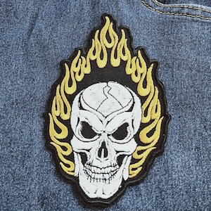 May include: Embroidered patch featuring a white skull with black eyes and a detailed jawline, surrounded by yellow flames on a black background. The patch is likely designed to be ironed or sewn onto clothing or accessories.