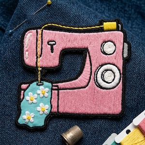 May include: A pink embroidered sewing machine patch with black outlining and yellow thread details. The patch includes a small blue tag with white daisy flowers. The patch is on a blue denim background.