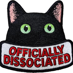 May include: Embroidered patch featuring a black cat face with green eyes and pink inner ears. The patch has a white border with the words "OFFICIALLY DISSOCIATED" in red lettering.