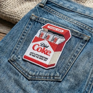 May include: Embroidered patch featuring a Diet Coke cigarette pack design. The patch is red, white, and black, with the text "Fridge Cigs" and "Just a little something to take the edge off." The patch is on a blue denim pocket.