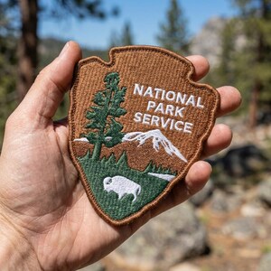 May include: A brown, shield-shaped embroidered patch featuring the text NATIONAL PARK SERVICE in white. The design includes a green pine tree, a white mountain peak, and a white bison silhouette against a brown background with a brown border.