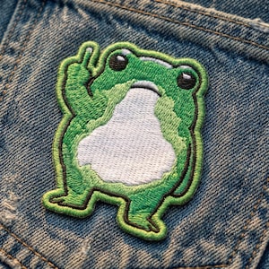 May include: Embroidered patch featuring a green frog giving the middle finger. The frog has a white belly, black eyes, and a green outline. The patch is sewn onto a denim jacket.