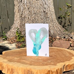 Puede incluir: Una tarjeta de felicitación blanca con un diseño de corazón abstracto en tonos plateados y turquesas. La obra de arte se muestra sobre una superficie de madera, con un tronco de árbol al fondo. Una pequeña firma es visible en la esquina inferior derecha.