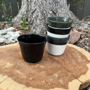 May include: A collection of handmade ceramic cups in various colors, including black, white, and green. The cups are stacked on a wooden surface, showcasing their unique shapes and textures. The cups are ideal for serving beverages.