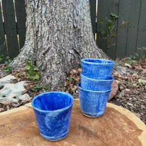 May include: Four handmade blue ceramic cups with a textured, marbled appearance. One cup is separate, while the other three are stacked. The cups are displayed on a natural wood surface, with a tree trunk and green foliage in the background.