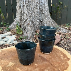 May include: Four handmade ceramic cups with a dark blue glaze and a white rim are arranged on a wooden surface. The cups have a slightly irregular shape, adding to their handcrafted appeal. The background features a tree trunk and a wooden fence.