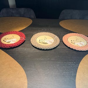 Könnte beinhalten: Drei handgefertigte Untersetzer mit einem Kaffeetassen-Design. Jeder Untersetzer hat eine Holzmitte mit einem gehäkelten Rand in Rot, Beige und Rosa. Die Untersetzer sind auf einem dunklen Holztisch platziert.