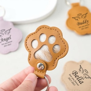 Puede incluir: Un llavero de cuero marrón con forma de huella de pata con un llavero plateado. La huella de la pata tiene un centro de piel blanca y marrón. Otros llaveros en el fondo tienen alas de ángel y los nombres "Angel", "Buddy" y "Bella" con fechas y la frase "love you forever".