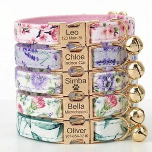 May include: A stack of five floral cat collars with gold-colored buckles and bells. Each collar features a different floral pattern in shades of pink, purple, and green. The buckles are engraved with names and contact information.