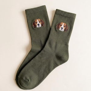 May include: A pair of olive green crew socks featuring an embroidered beagle dog face on the upper ribbed section of each sock. The beagle embroidery displays brown, white, and black fur details with a distinct white stripe on the snout.