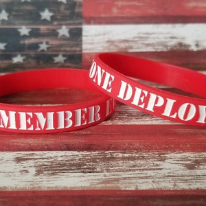 May include: Two red silicone wristbands with white lettering. One reads "REMEMBER THEM" and the other reads "ONE DEPLOYED". The background is a wooden surface with a painted American flag.