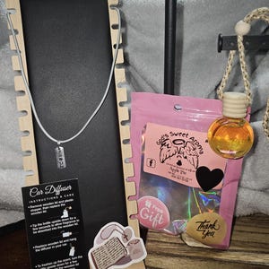 May include: A display featuring a silver necklace with a "MIRACLE" pendant, a car air freshener, and a pink bag with a "God's Sweet Aroma" label. The car air freshener is a small glass bottle with a wooden lid. A sticker reads "Thank You".