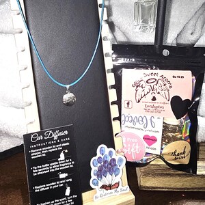 May include: A display featuring a blue cord necklace with a silver pendant, a car air freshener, and a small glass bottle. Also included are stickers and a black bag with text that reads "Sweet Aroma".