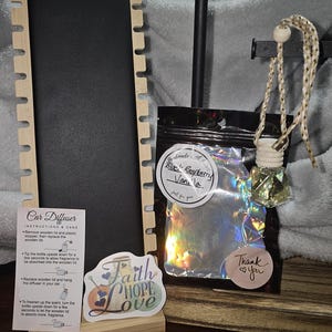 Puede incluir: Un ambientador para coche con aroma a vainilla y frambuesa negra, expuesto con un soporte de madera y una pegatina decorativa que dice "Faith Hope Love". El producto incluye instrucciones y una pegatina de agradecimiento.