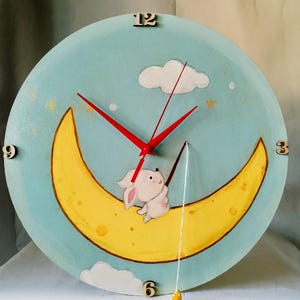 Puede incluir: Un reloj de pared redondo pintado a mano con un fondo azul claro. El reloj presenta una luna creciente amarilla con un conejito blanco, manecillas rojas y números de madera. Una estrella amarilla cuelga debajo del reloj.