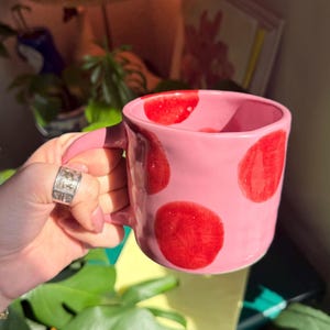 May include: A pink ceramic mug with large, irregular red polka dots. The mug has a rounded handle and a smooth, glossy finish. The mug is held by a hand with a silver ring.