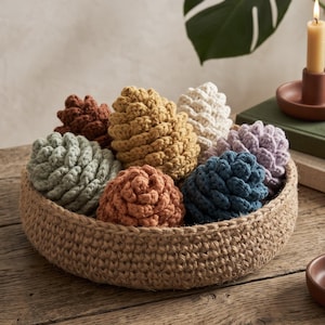May include: A round, woven jute basket filled with several crocheted pinecone-shaped ornaments in various colors, including mustard yellow, sage green, terracotta, cream, dusty blue, and soft lavender, arranged on a wooden surface.
